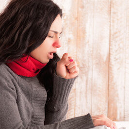 Coughing beauty. Side view image of young sick woman with scarf on her neck sitting in bed and coughing with her country house on the background