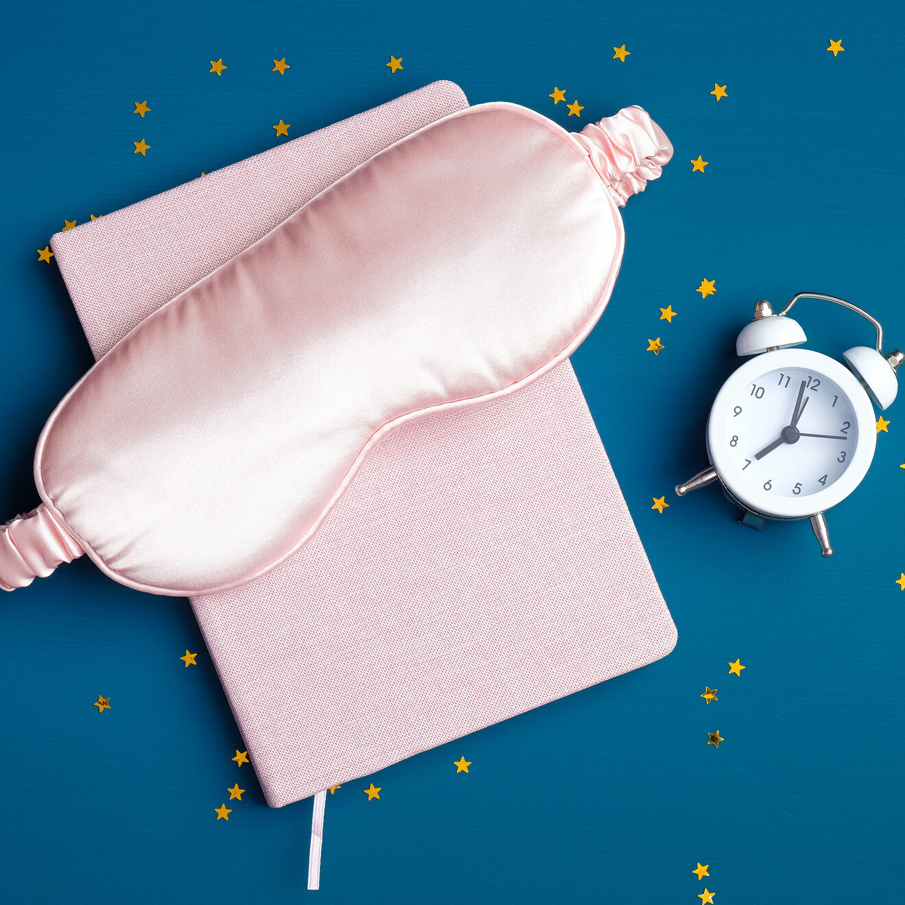 Good night and healthy sleeping concept. Top view sleeping accessories on dark blue background. Flat lay composition with sleep eye mask, pink dream book, alarm clock, confetti. Insomnia treatment.