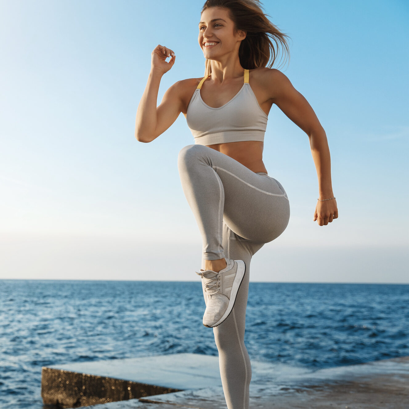 Motivated happy sporty woman wearing sports bra, sneakers enjoying excercise, training outdoors near sea, workout quay, jogging, running, jumping energized, smiling during productive fitness session.